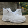A pair of white high-top sneakers, identified as Air Jordan 12 models, shown outdoors on a stone surface with a blurred background of greenery and a wall.