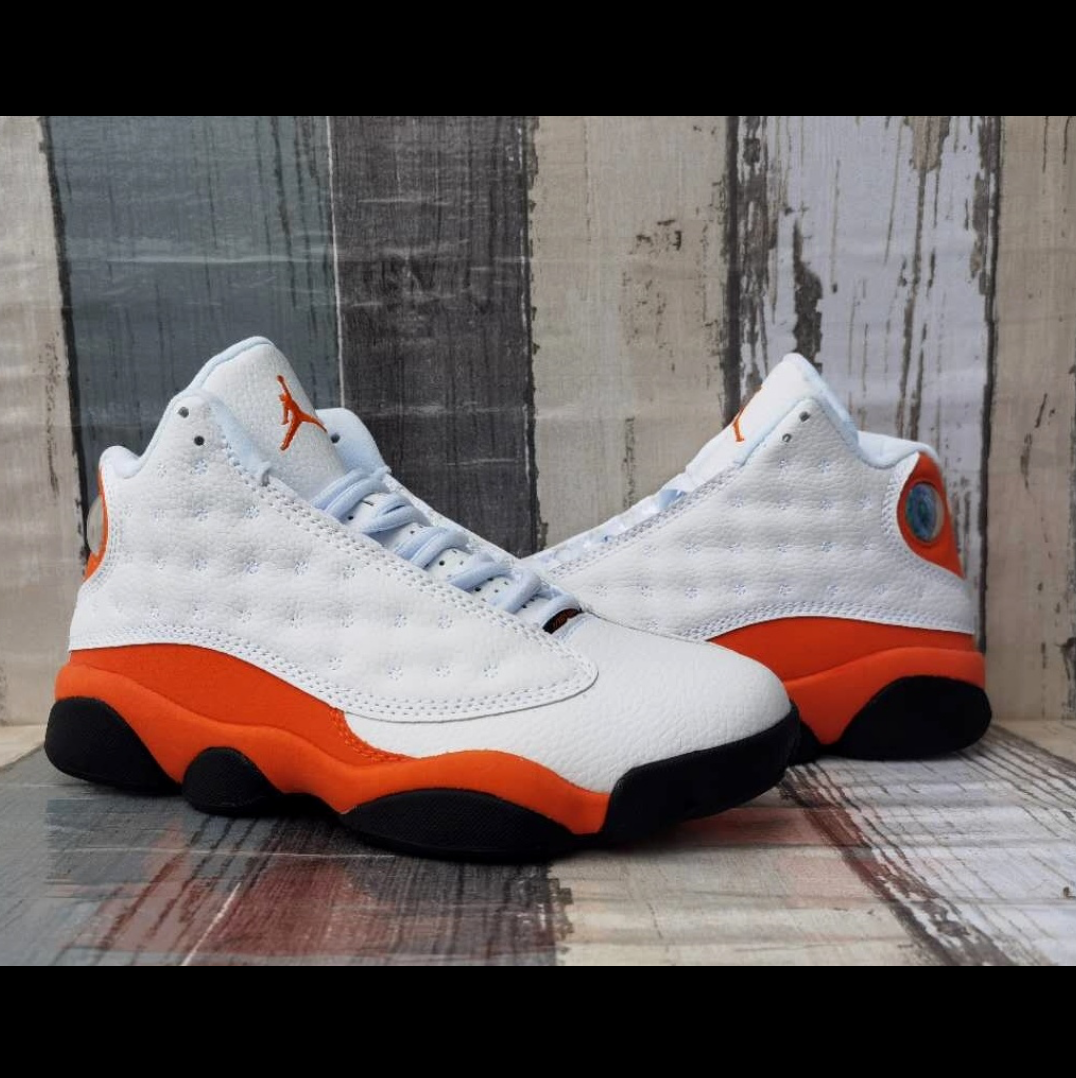 A pair of white and orange Air Jordan 13 sneakers displayed on a rustic wooden surface, with a smartphone screenshot interface showing the product photo and text details.